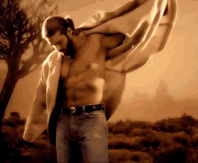 a shirtless man with a ponytail is standing in a field with a tree in the background