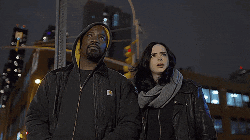 a man in a carhartt jacket stands next to a woman in a scarf and a man in a red mask