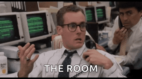 a man in a suit and tie is talking on a phone and the room is written on the screen behind him .