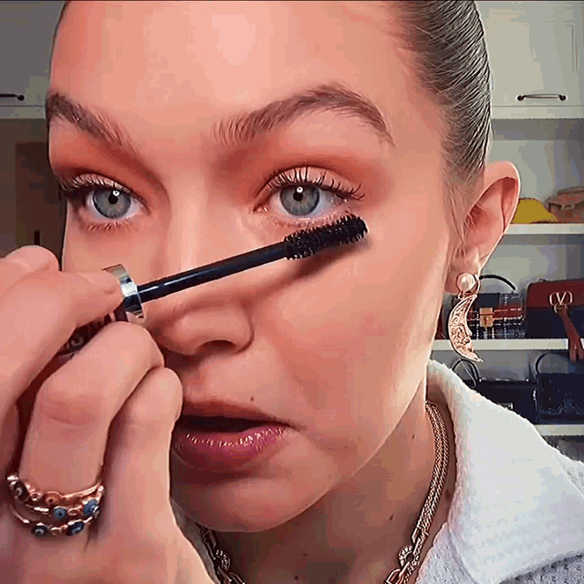 a woman is applying mascara to her eye while wearing a necklace with the letter v on it