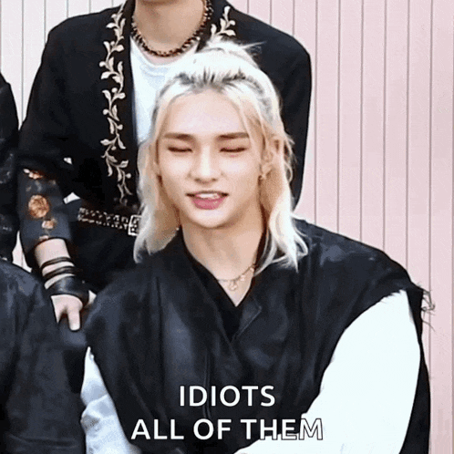 a man with blonde hair is sitting in front of a group of people and says idiots all of them