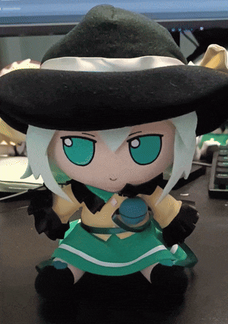 a stuffed doll with green eyes and a black hat sits on a desk in front of a computer monitor