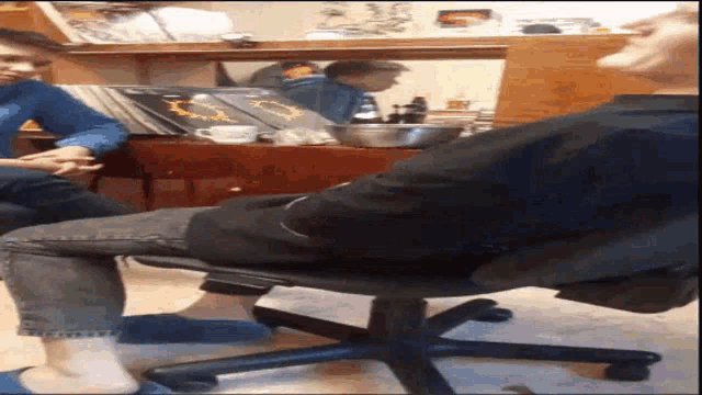 a man is sitting in an office chair with his legs crossed while a woman sits at a table .