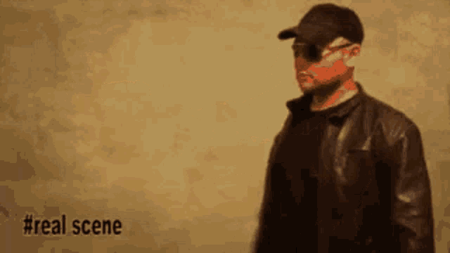 a man wearing sunglasses and a hat is standing in front of a wall with #real scene written below him
