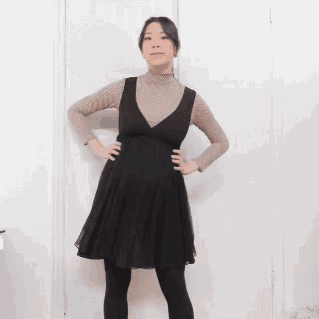 a woman wearing a black dress and a tan turtleneck is standing in front of a white wall .