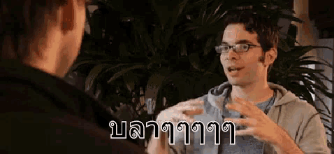 a man with glasses is talking to another man in a room with a plant in the background .