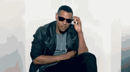a man wearing sunglasses and a leather jacket is sitting down and talking on a cell phone .