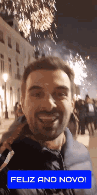 a man is smiling in front of a fireworks display and a feliz ano novo sticker