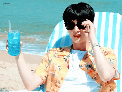 a man wearing sunglasses sits on a beach holding a blue drink with a straw