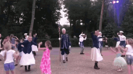 a group of children are dancing in a park with a man in a blue suit holding a sword .