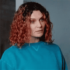 a woman with red curly hair is wearing a blue sweater and looking at the camera .