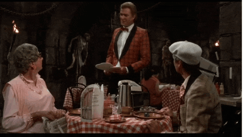 a man in a tuxedo is holding a plate and talking to two women at a table in a restaurant