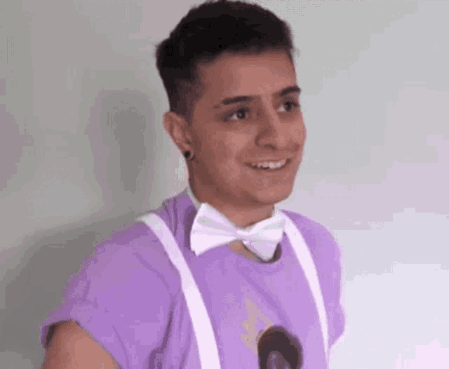 a young man wearing a purple shirt , white suspenders and a bow tie is smiling .