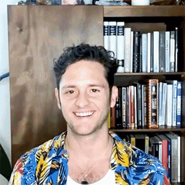 a man in a hawaiian shirt is smiling in front of a bookshelf filled with books .