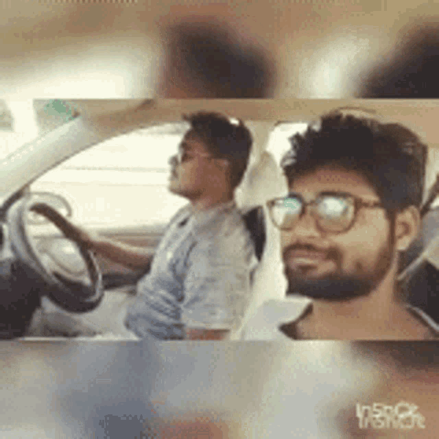a man with glasses is driving a car next to another man with a beard .