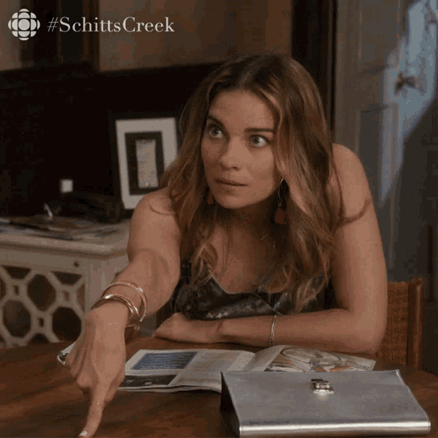 a woman sitting at a table with a #schitts creek logo on the bottom