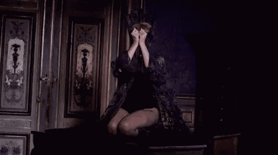 a woman in a black dress is kneeling down in a dark room with a mask on her face .