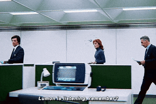 a man in a suit is talking to a woman in an office cubicle while another man reads a book