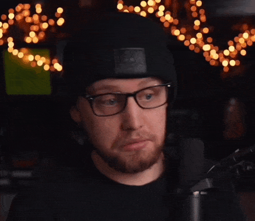 a man wearing glasses and a beanie looks at the camera with christmas lights in the background