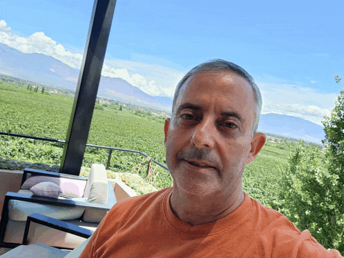 a man in an orange shirt takes a selfie with a view of a vineyard in the background