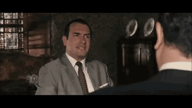 a man in a suit and tie is talking to another man in a dark room