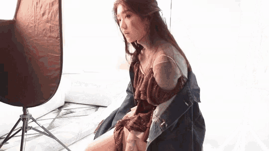 a woman in a dress and denim jacket is sitting on the floor in front of a tripod .