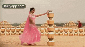 a woman in a pink dress is dancing in front of a row of pots on a sandy beach .