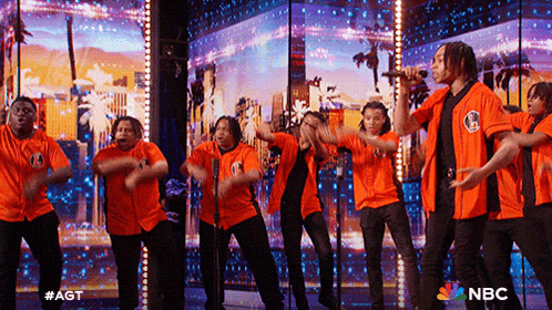 a group of men in orange shirts are performing on a stage with a nbc logo in the background