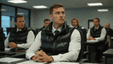 a man in a black vest sits at a desk in a classroom