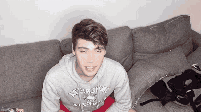 a young man wearing a grey hoodie that says ' a' on it sits on a couch