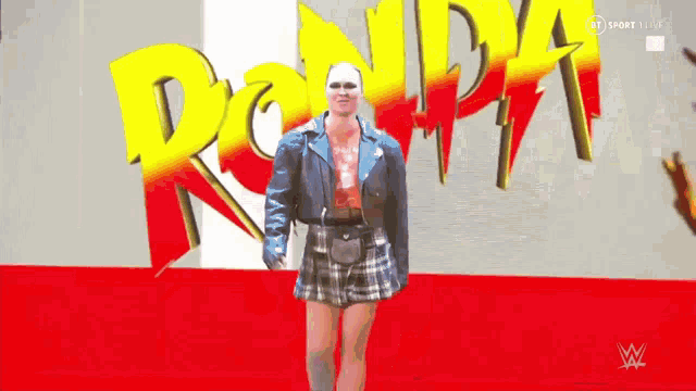 a woman in a blue jacket and plaid skirt stands in front of a sign that says ronda