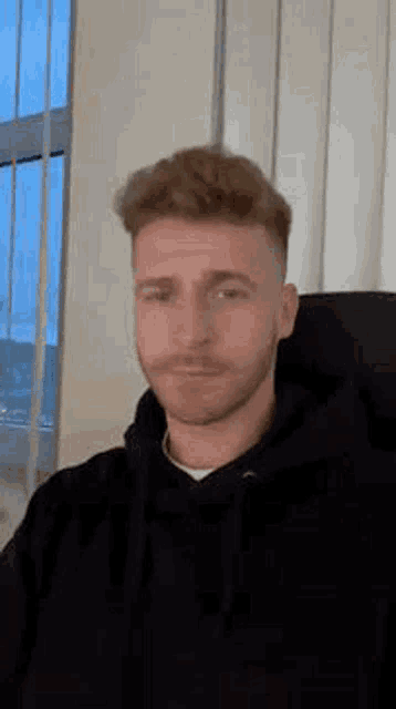 a man with a beard is wearing a black hoodie and sitting in a chair .
