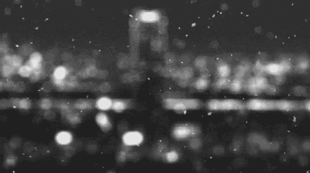 a black and white photo of snow falling on a city at night