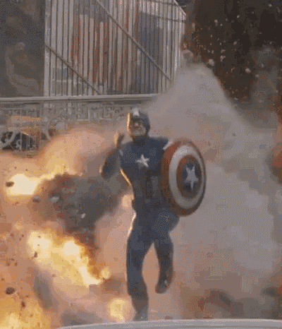 captain america is running through a fire while holding a shield in front of an explosion .
