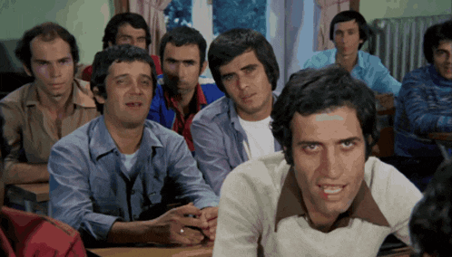 a group of men are sitting in a classroom and one of them is wearing a ring on his finger