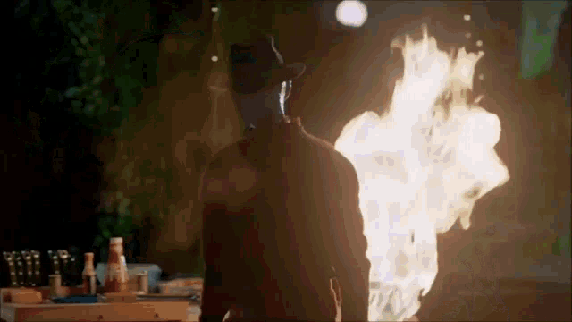 a man in a hat is standing in front of a burning fire .