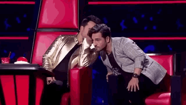 two men are sitting next to each other in red chairs and one of them is whispering into the other 's ear .