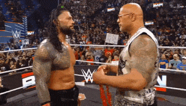 roman reigns and the rock are standing in a wrestling ring holding hands .