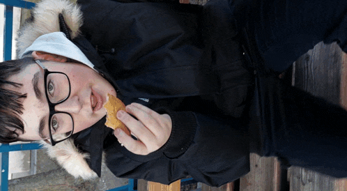 a boy wearing glasses and a black jacket is eating a piece of food