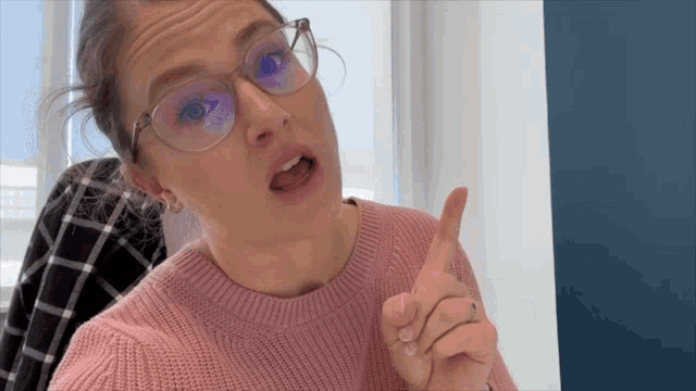 a woman wearing glasses and a pink sweater holds up her finger