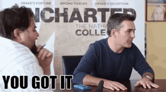 two men are sitting at a table in front of a sign that says uncharted