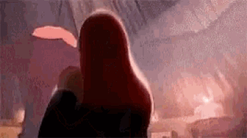 a woman with long red hair is standing in front of a fire in a dark room .