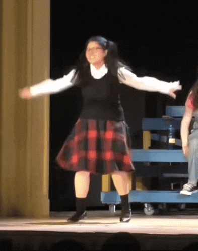 a girl in a plaid skirt is dancing on a stage with her arms outstretched