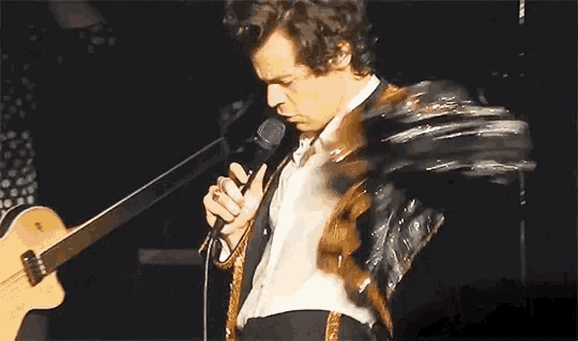 harry styles is singing into a microphone while holding a guitar .