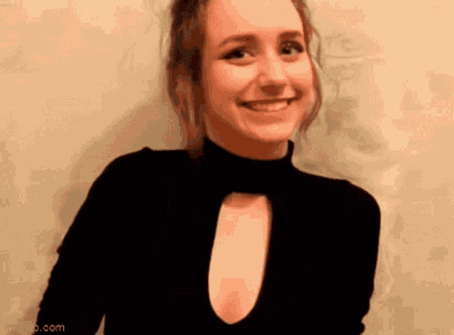 a woman in a black turtleneck is smiling and standing in front of a white wall .