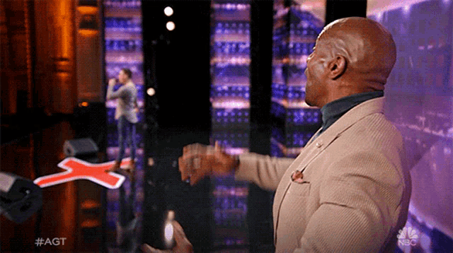 a man in a tan suit stands in front of a red cross on a stage with #agt written on it