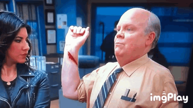 a man in a tan shirt and tie has a wound on his arm and a woman in a leather jacket looks on