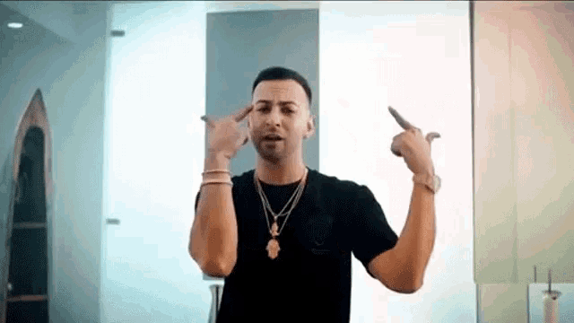 a man wearing a black shirt and a gold necklace is pointing at himself .