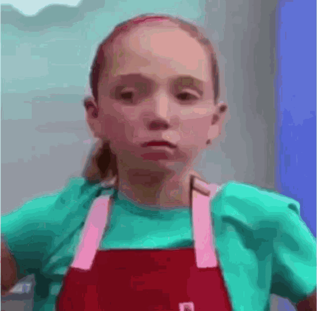 a young girl wearing an apron and a green shirt is making a face .