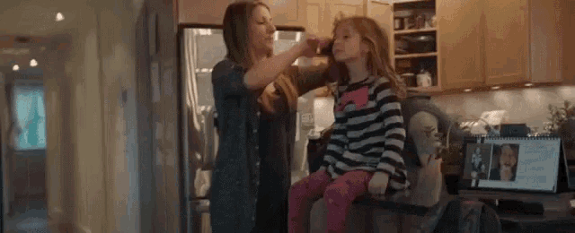 a woman is brushing a little girl 's hair in a kitchen in front of a laptop .